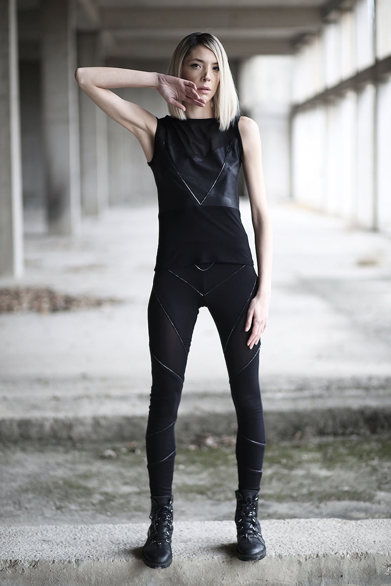 Op de afbeelding: Een vrouw draagt een zwart tanktop met een lederen paneel en zwarte leggings met geometrische stiksels. Ze draagt zwarte combatlaarzen en staat in een betonnen gebouw.