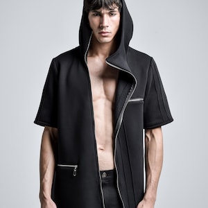 Puede incluir: Chaqueta negra con capucha y manga corta con cierre de cremallera. La chaqueta presenta un dise&ntilde;o a medida con costuras verticales y una cremallera frontal que se extiende hasta la cintura. El modelo lleva pantalones negros.