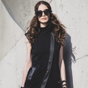 May include: A woman wearing a black sleeveless dress with a high collar and a diagonal zipper detail. She is also wearing round black sunglasses and leather accessories, including wrist cuffs and a thigh garter. The background is a textured concrete wall.