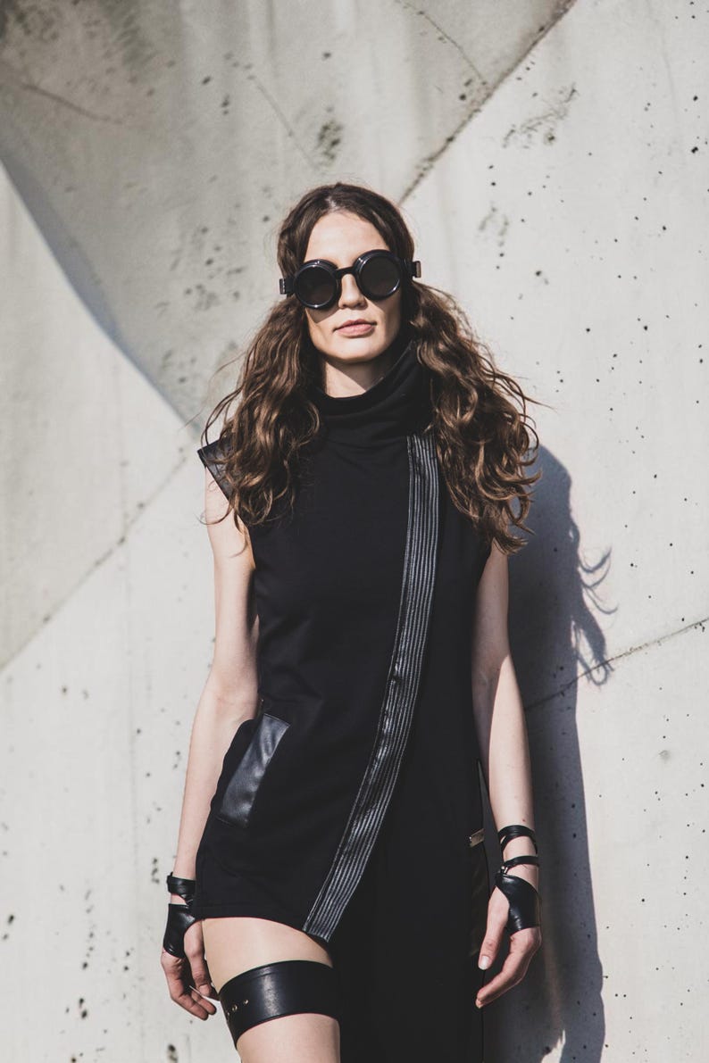 May include: A woman wearing a black sleeveless dress with a high collar and a diagonal zipper detail. She is also wearing round black sunglasses and leather accessories, including wrist cuffs and a thigh garter. The background is a textured concrete wall.