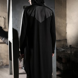 May include: Black hooded long-sleeve shirt with a unique asymmetrical hem. The shirt features a textured panel on the upper back and shoulders. The model is wearing black shorts and boots. The clothing is a dark, modern style.