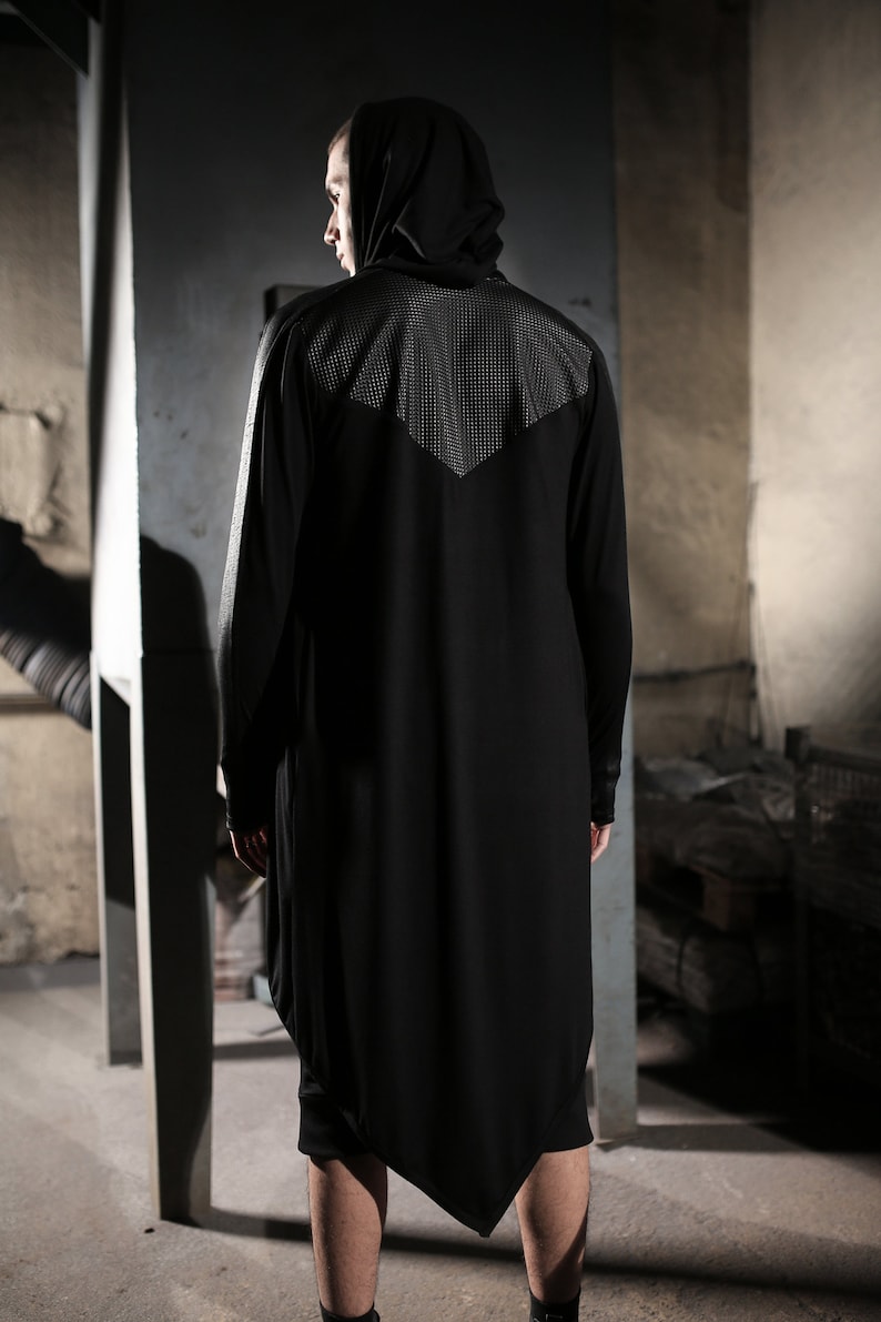 May include: Black hooded long-sleeve shirt with a unique asymmetrical hem. The shirt features a textured panel on the upper back and shoulders. The model is wearing black shorts and boots. The clothing is a dark, modern style.