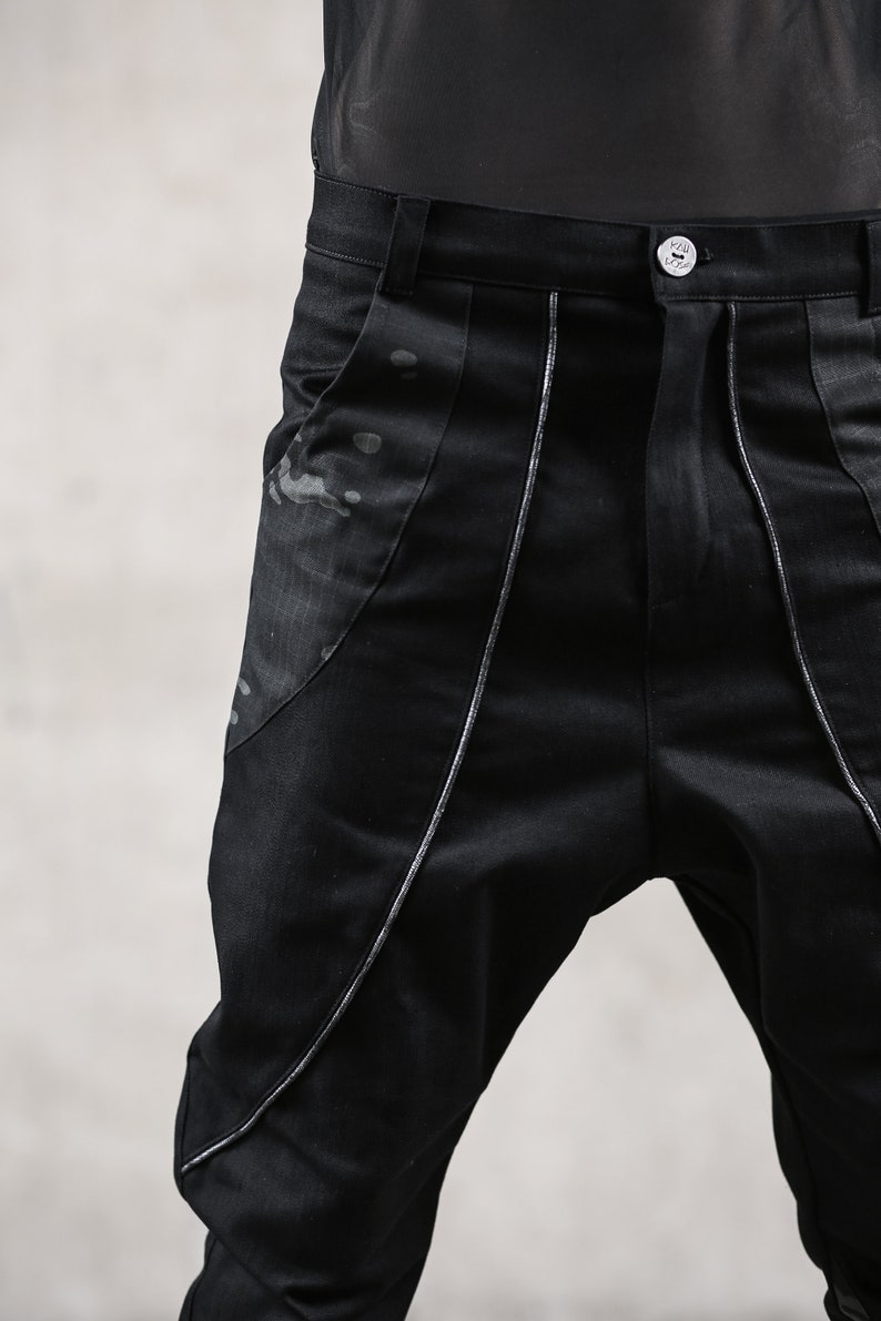 May include: Black pants with a unique design. The pants feature silver stitching along the seams and a decorative panel on the front. The fabric has a subtle sheen, and the pants have a button closure.