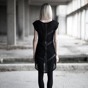 May include: A black dress with a sheer overlay and leather-like accents. The dress features a unique design with geometric lines and is paired with black leggings and boots. The image is taken in a concrete industrial setting.