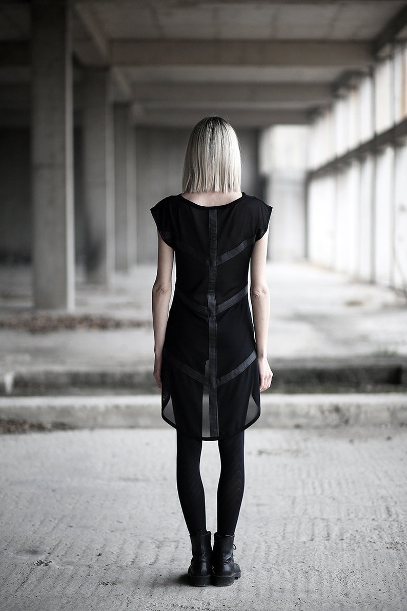 May include: A black dress with a sheer overlay and leather-like accents. The dress features a unique design with geometric lines and is paired with black leggings and boots. The image is taken in a concrete industrial setting.