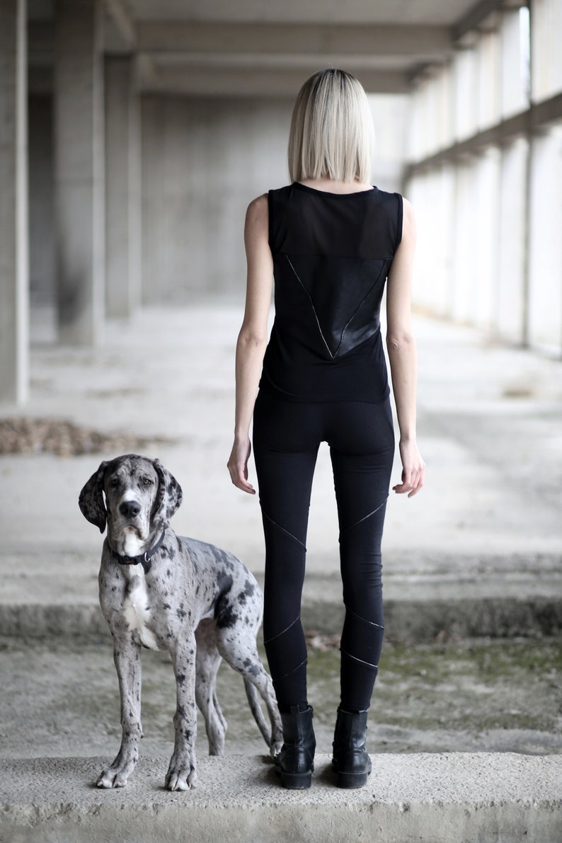 Op de afbeelding: Een vrouw met blond haar draagt een zwart, mouwloos topje en een zwarte legging met een grijze en witte Great Dane die naast haar staat in een betonnen gebouw.