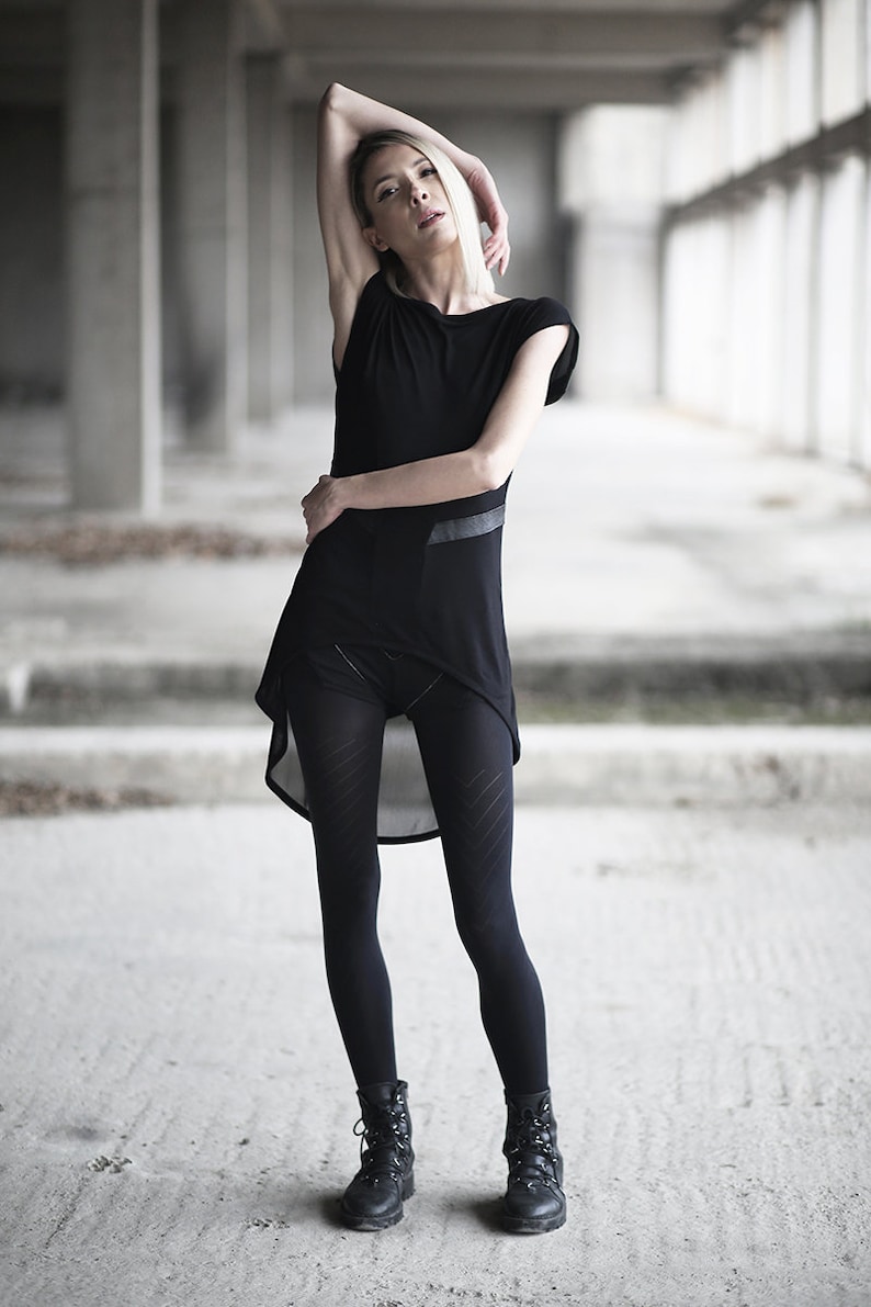 May include: A black outfit consisting of a short-sleeved top with a layered asymmetrical hem, black leggings with a geometric pattern, and black combat boots. The outfit is worn in a concrete industrial setting.