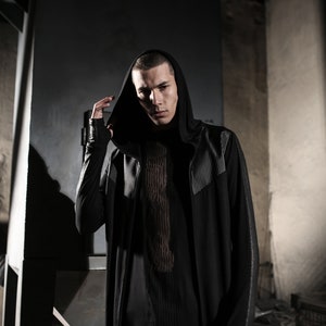May include: A black hooded long-sleeve top with a textured design on the shoulders and sleeves. The top is layered over a black shirt with a vertical striped pattern. The model is wearing a ring.