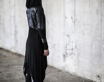 May include: A black hooded tunic featuring a textured dark grey upper back panel, long sleeves with ribbed cuffs, and an asymmetrical hemline that is longer in the back. The garment is paired with black trousers and black leather lace-up boots.