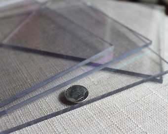 Puede incluir: Tres láminas de acrílico transparente apiladas una encima de la otra sobre una superficie de tela marrón claro. Una moneda de diez céntimos de euro plateada se coloca en la lámina inferior.