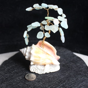 Peut inclure: Une sculpture d'arbre en verre de mer avec une base en coquillage. L'arbre est fait de fil de fer et de verre de mer, et la base est un grand coquillage avec une base en corail blanc.