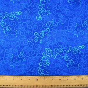 May include: Close-up of a blue fabric with a swirling, scroll-like pattern in varying shades of blue and turquoise. A wooden ruler with inch markings is at the bottom of the image, indicating the fabric's potential for crafting or sewing projects.