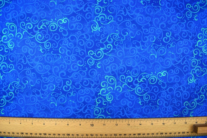 May include: Close-up of a blue fabric with a swirling, scroll-like pattern in varying shades of blue and turquoise. A wooden ruler with inch markings is at the bottom of the image, indicating the fabric's potential for crafting or sewing projects.