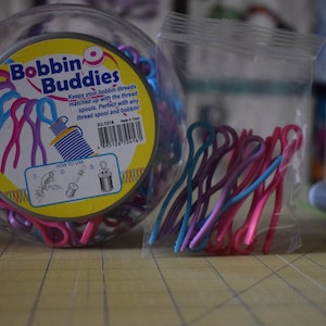 May include: A clear plastic jar filled with colourful bobbin buddies, which are plastic thread holders. The jar has a yellow label with the text "Bobbin Buddies" and instructions on how to use the product.