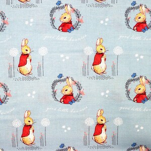 May include: A blue fabric with a repeating pattern of a yellow rabbit wearing a red jacket. The rabbit is surrounded by a wreath of green leaves and white flowers. The text "good little bunnies" is printed below the rabbit.