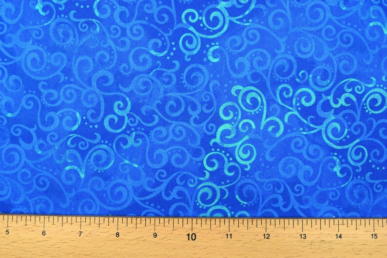 May include: Close-up of a blue fabric with a swirling, scroll-like pattern in shades of blue and teal. The fabric is measured with a wooden ruler marked in inches. This textile is suitable for various craft projects.