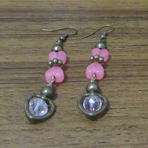 Puede incluir: Un par de pendientes colgantes con cuentas de corazón rosa y detalles de cristal transparente. Los pendientes tienen un acabado bronce.