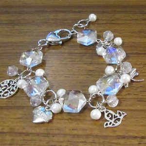 May include: A silver charm bracelet featuring clear crystal beads, silver charms, and white pearl accents. The charms include a tree, a heart with the word "love" on it, and a dove.