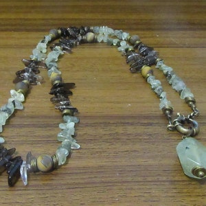 Op de afbeelding: Een lange ketting gemaakt van bruine, groene en heldere stenen met een grote, lichtgroene stenen hanger. De ketting heeft een messing sluiting.