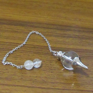 Può includere: Un pendolo di cristallo trasparente con una catena d'argento e due perle di cristallo bianche. Il pendolo è su una superficie di legno.