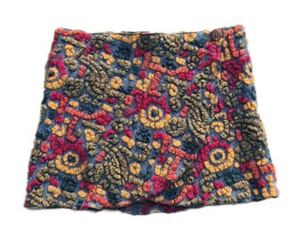May include: A colorful, embroidered wrap skirt. The skirt features a blue background with intricate patterns in red, yellow, and green. A single button closure is visible. The skirt is a unique piece of clothing.