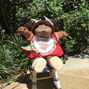 Puede incluir: Una muñeca Cabbage Patch Kid de color marrón con el pelo marrón en coletas que lleva un vestido rojo con ribete de encaje blanco y un babero blanco con una jirafa marrón bordada. La muñeca está sentada en un taburete pintado de blanco y azul.