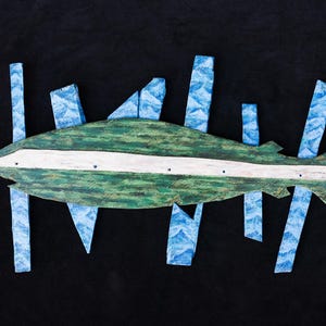 May include: Wooden fish wall art with a green body and white stripe, accented by blue vertical and angled elements. The artwork is set against a black background, creating a striking contrast. The fish design has a rustic, hand-painted appearance.