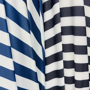 K&ouml;nnte beinhalten: Nahaufnahme von zwei Stoffen mit kr&auml;ftigen, horizontalen Streifen. Ein Stoff hat marineblaue und wei&szlig;e Streifen, w&auml;hrend der andere schwarz-wei&szlig;e Streifen hat. Die Stoffe sind drapiert und erzeugen weiche Falten und Schatten.