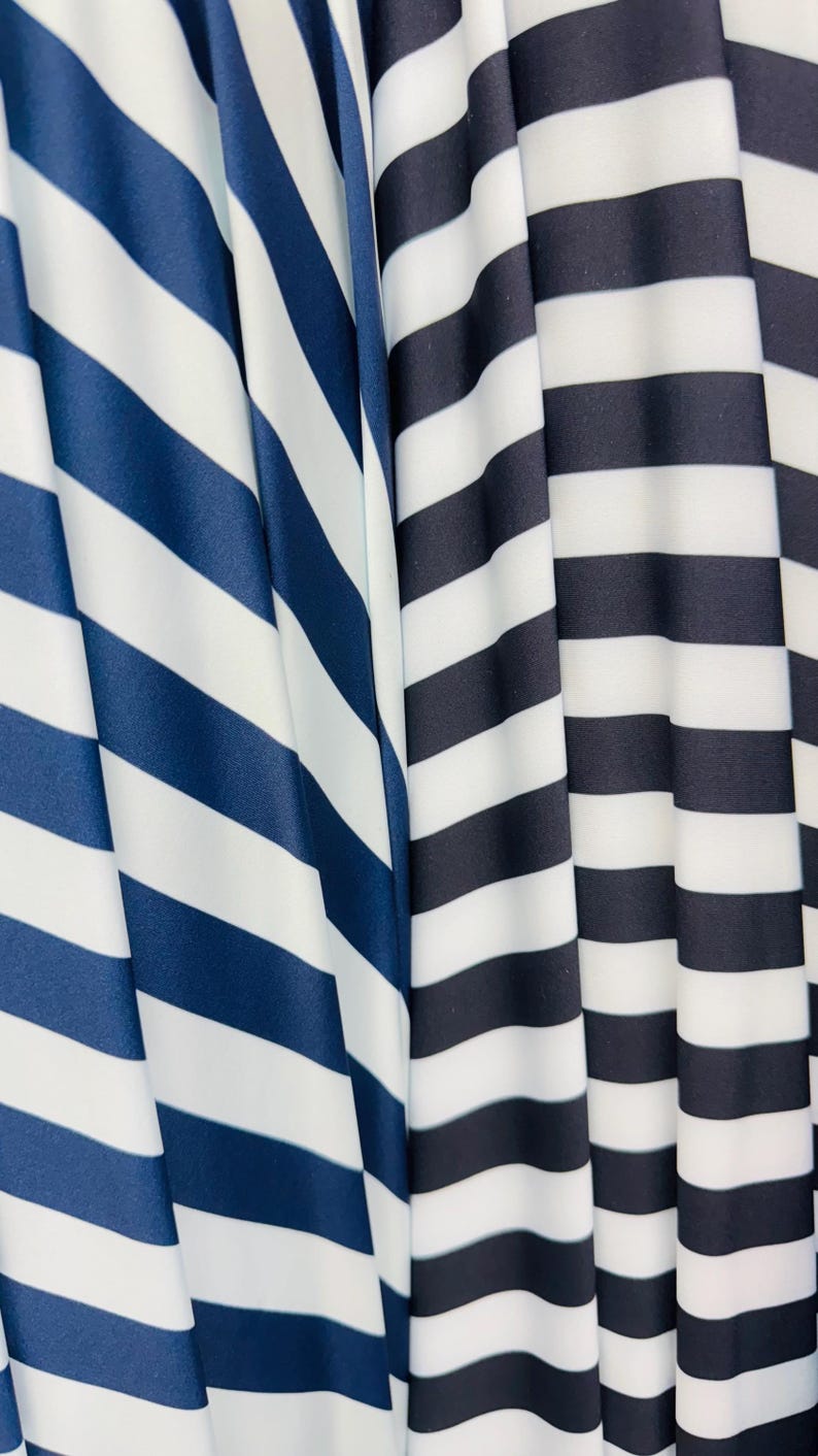 K&ouml;nnte beinhalten: Nahaufnahme von zwei Stoffen mit kr&auml;ftigen, horizontalen Streifen. Ein Stoff hat marineblaue und wei&szlig;e Streifen, w&auml;hrend der andere schwarz-wei&szlig;e Streifen hat. Die Stoffe sind drapiert und erzeugen weiche Falten und Schatten.