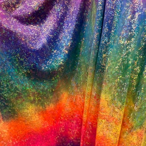 K&ouml;nnte beinhalten: Ein regenbogenfarbener, schimmernder Stoff mit einer gesprenkelten Textur. Der Stoff ist gefaltet und zeigt einen Farbverlauf von Lila bis Gelb.