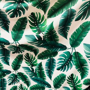 Op de afbeelding: Een witte stof met een herhalend patroon van groene tropische bladeren. De bladeren zijn in verschillende vormen en maten, waaronder grote monstera bladeren en kleinere palmbladeren.