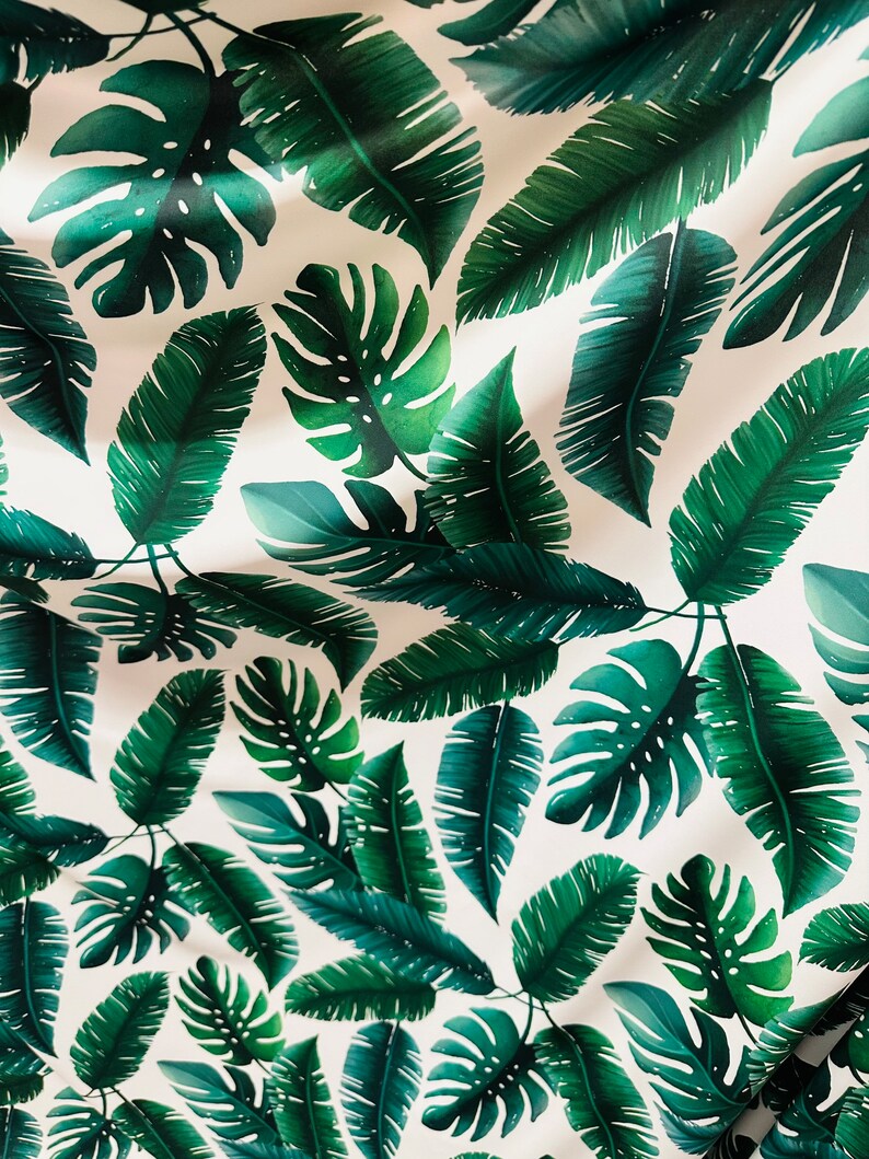 Op de afbeelding: Een witte stof met een herhalend patroon van groene tropische bladeren. De bladeren zijn in verschillende vormen en maten, waaronder grote monstera bladeren en kleinere palmbladeren.