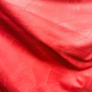 Könnte beinhalten: Nahaufnahme eines leuchtend roten Ledermaterials mit geometrischem Muster. Das Leder hat eine strukturierte Oberfläche und ist gefaltet, wodurch Schatten und Lichtreflexe entstehen, die das Design hervorheben. Der Gesamteindruck ist reichhaltig und taktil.