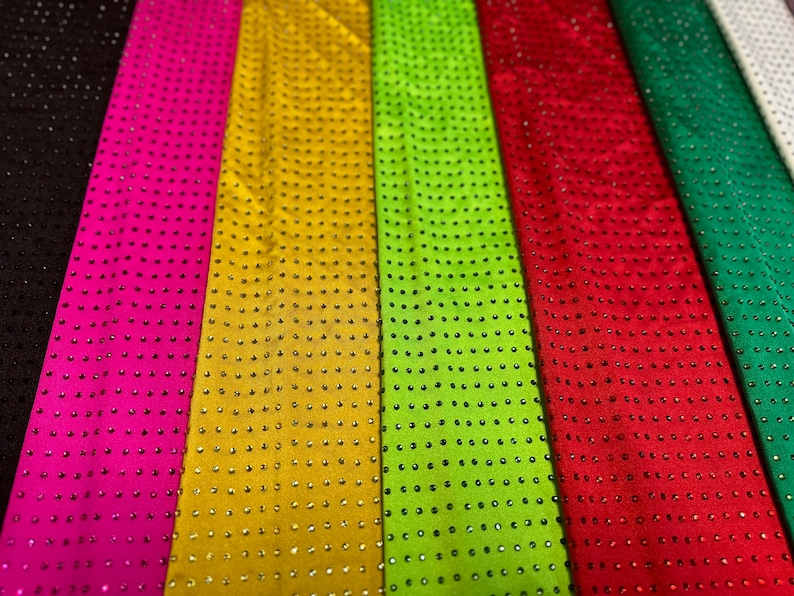 Op de afbeelding: Een collectie verticale stroken stof in zwart, felroze, goud, limoengroen, rood, smaragdgroen en wit. Elke strook is voorzien van een uniform raster van kleine, reflecterende, ronde studs of strass-steentjes in consistente rijen en kolommen.
