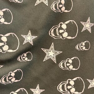 May include: Dark green fabric featuring a repeating pattern of white skulls and star shapes. The skulls have eye sockets and teeth, and the stars have a five-point design.
