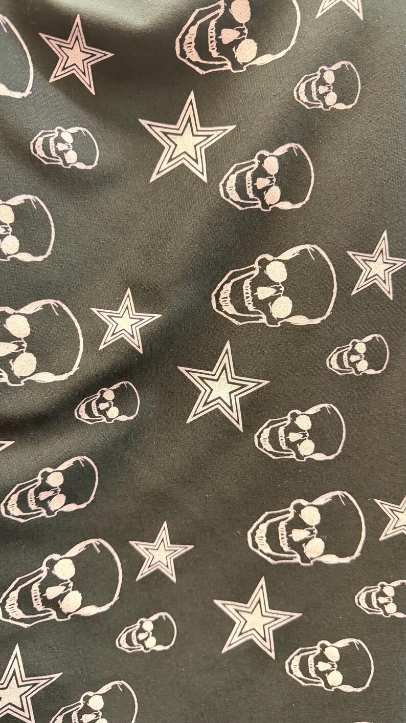 May include: Dark green fabric featuring a repeating pattern of white skulls and star shapes. The skulls have eye sockets and teeth, and the stars have a five-point design.