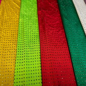 Op de afbeelding: Een collectie stoffen stroken die naast elkaar zijn gerangschikt in diverse kleuren, waaronder roze, goud, limoengroen, rood, donkergroen, wit en paars. Elke strook is voorzien van een uniform raster van kleine, reflecterende studs.