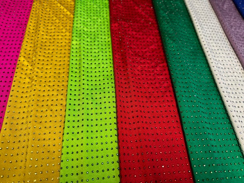 Op de afbeelding: Een collectie stoffen stroken die naast elkaar zijn gerangschikt in diverse kleuren, waaronder roze, goud, limoengroen, rood, donkergroen, wit en paars. Elke strook is voorzien van een uniform raster van kleine, reflecterende studs.