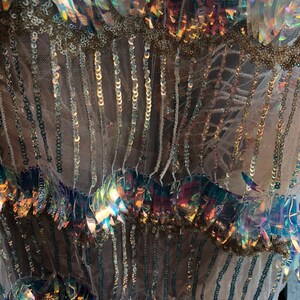 K&ouml;nnte beinhalten: Nahaufnahme von schimmernden Pailletten und Perlen, die auf einen durchsichtigen Stoff gen&auml;ht sind. Die Pailletten sind in Reihen angeordnet und sind eine Mischung aus Gold- und Regenbogenfarben.