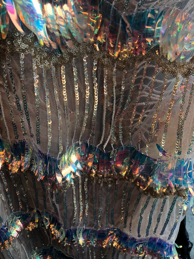 K&ouml;nnte beinhalten: Nahaufnahme von schimmernden Pailletten und Perlen, die auf einen durchsichtigen Stoff gen&auml;ht sind. Die Pailletten sind in Reihen angeordnet und sind eine Mischung aus Gold- und Regenbogenfarben.