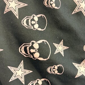 May include: Dark green fabric featuring a repeating pattern of white skulls and stars. The skulls have eye sockets, noses, and teeth. The stars are five-pointed.