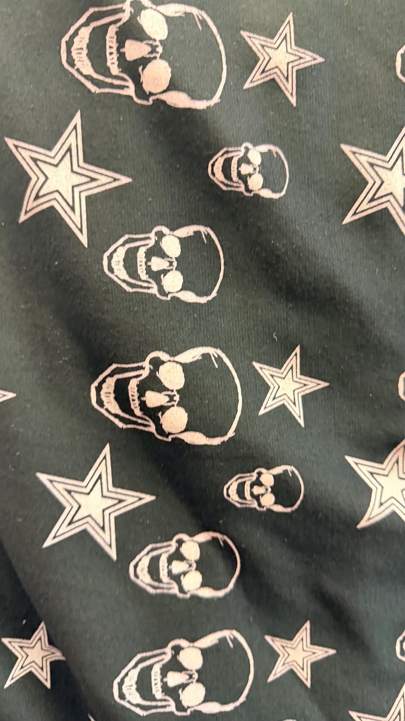 May include: Dark green fabric featuring a repeating pattern of white skulls and stars. The skulls have eye sockets, noses, and teeth. The stars are five-pointed.