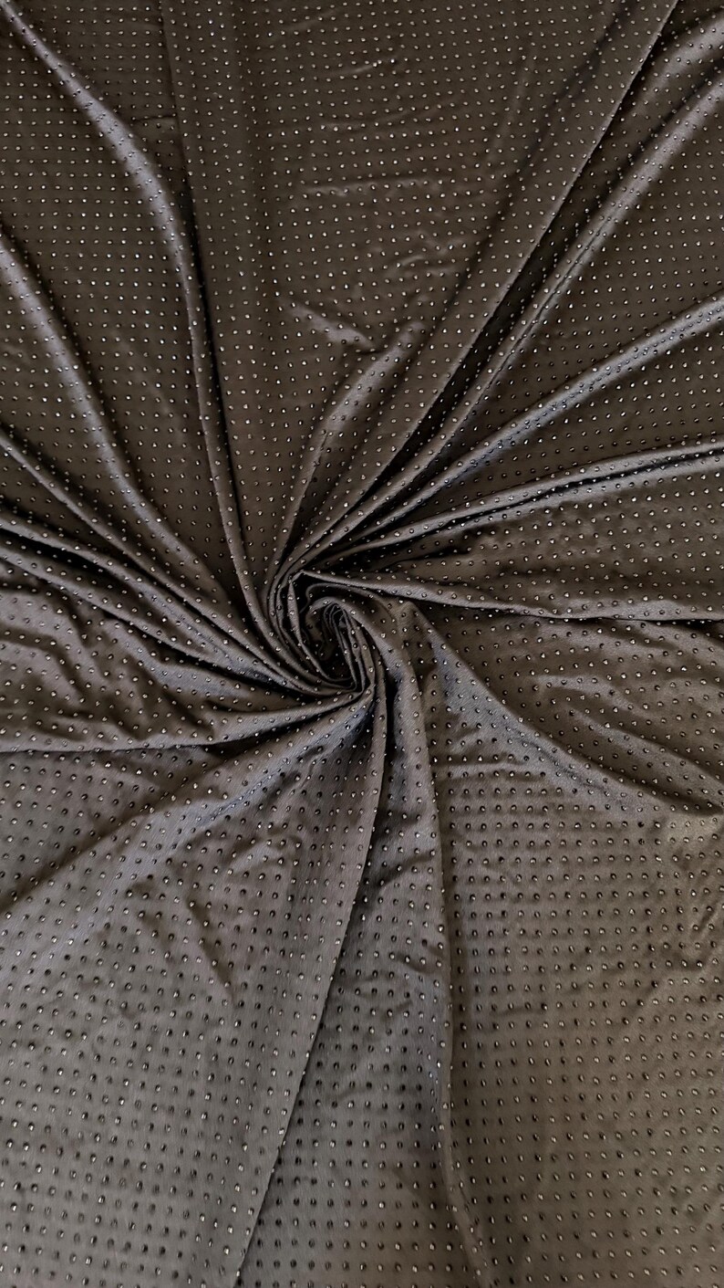 Op de afbeelding: Een donkerbruine stof met een herhalend patroon van kleine, verhoogde, cirkelvormige stippen over het gehele oppervlak. Het materiaal is in het midden samengebracht, waardoor diepe plooien en draperingen ontstaan die de textuur en de consistente afstand tussen de stippen benadrukken.