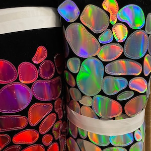 May include: Two rolls of black fabric with iridescent, sewn-on, oval-shaped patches. The fabric on the left has pink and red patches, while the fabric on the right has blue and green patches.