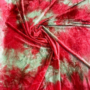 Op de afbeelding: Close-up van een stof met een tie-dye patroon. De stof is een mix van rode en lichtgroene kleuren, waarbij het rood het ontwerp domineert. De stof is gevouwen en gedrapeerd, waardoor een textuur ontstaat. De stof lijkt zacht en vloeiend.
