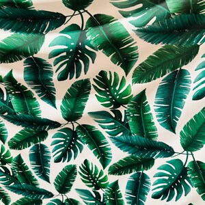 Op de afbeelding: Een witte stof met een groen tropisch bladmotief. De stof is gevouwen en de bladeren zijn in verschillende maten en vormen.