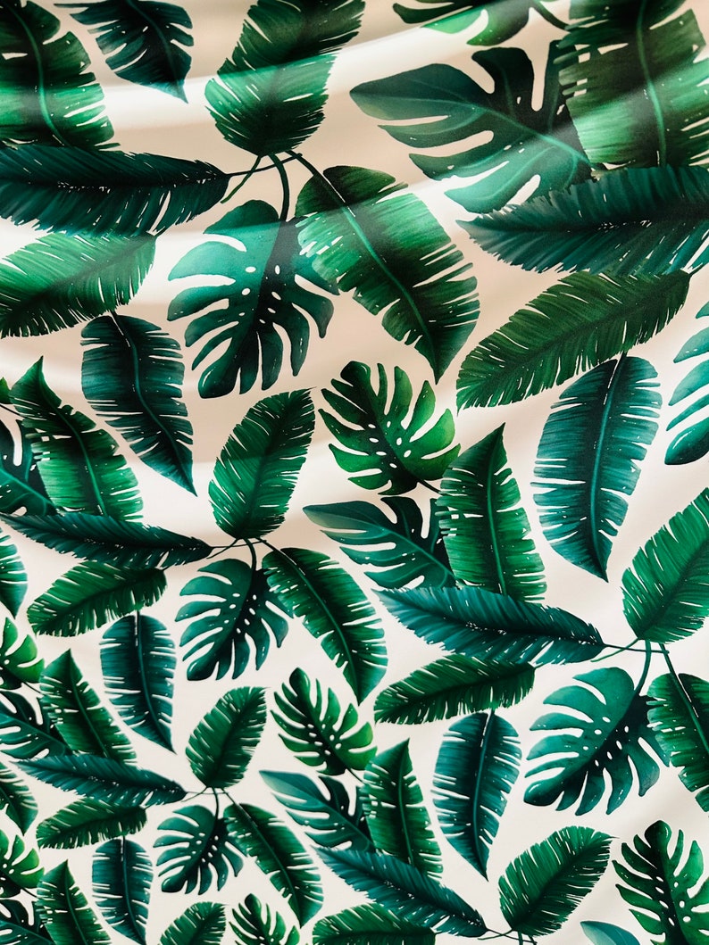Op de afbeelding: Een witte stof met een groen tropisch bladmotief. De stof is gevouwen en de bladeren zijn in verschillende maten en vormen.