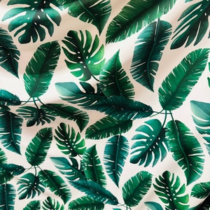Op de afbeelding: Witte stof met een herhalend patroon van groene tropische bladeren. De bladeren zijn in verschillende maten en vormen, waaronder grote monstera bladeren en kleinere palmbladeren.