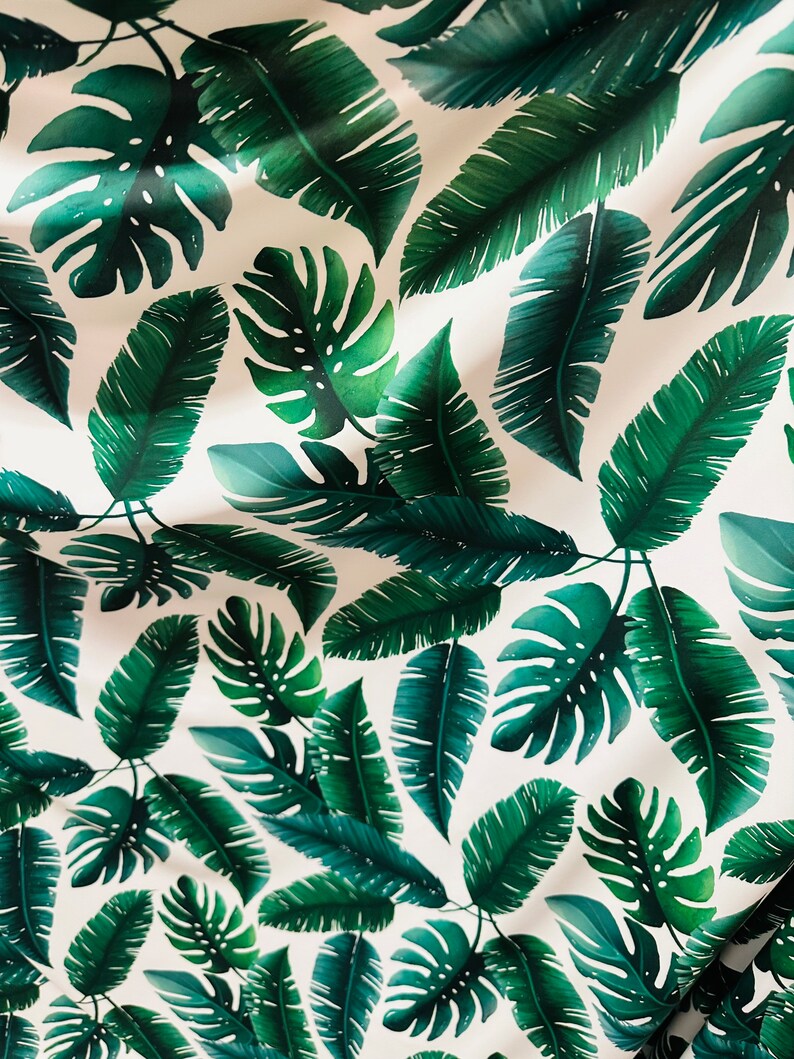 Op de afbeelding: Witte stof met een herhalend patroon van groene tropische bladeren. De bladeren zijn in verschillende maten en vormen, waaronder grote monstera bladeren en kleinere palmbladeren.
