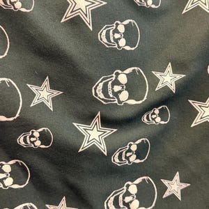 May include: Dark green fabric featuring a repeating pattern of white skulls and five-pointed stars. The skulls have eye sockets and teeth, and the stars have a double outline.
